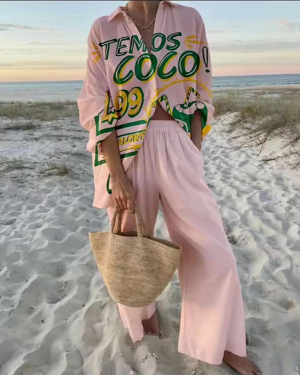 Woman wearing pink Savanna Siren bikini with matching cover-up on beach at sunset holding straw bag.