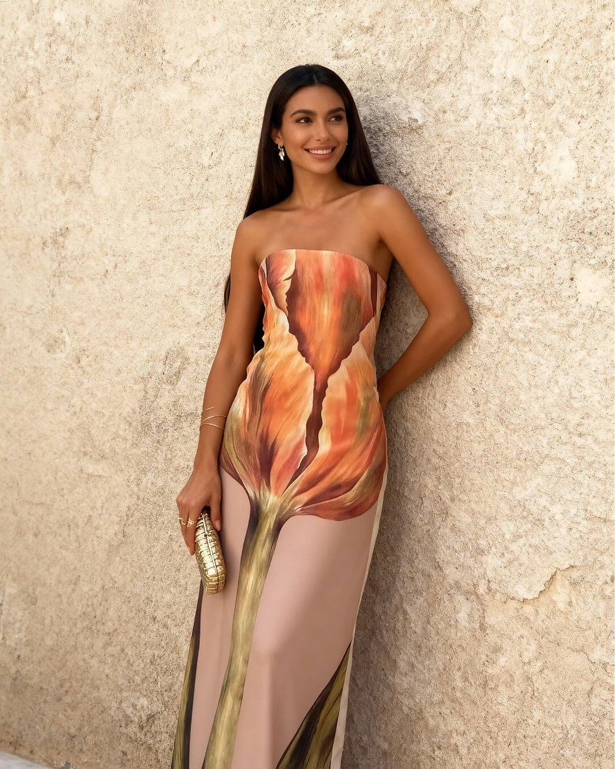 Woman wearing strapless Maison Amara dress with coral and gold floral print design against textured beige wall.