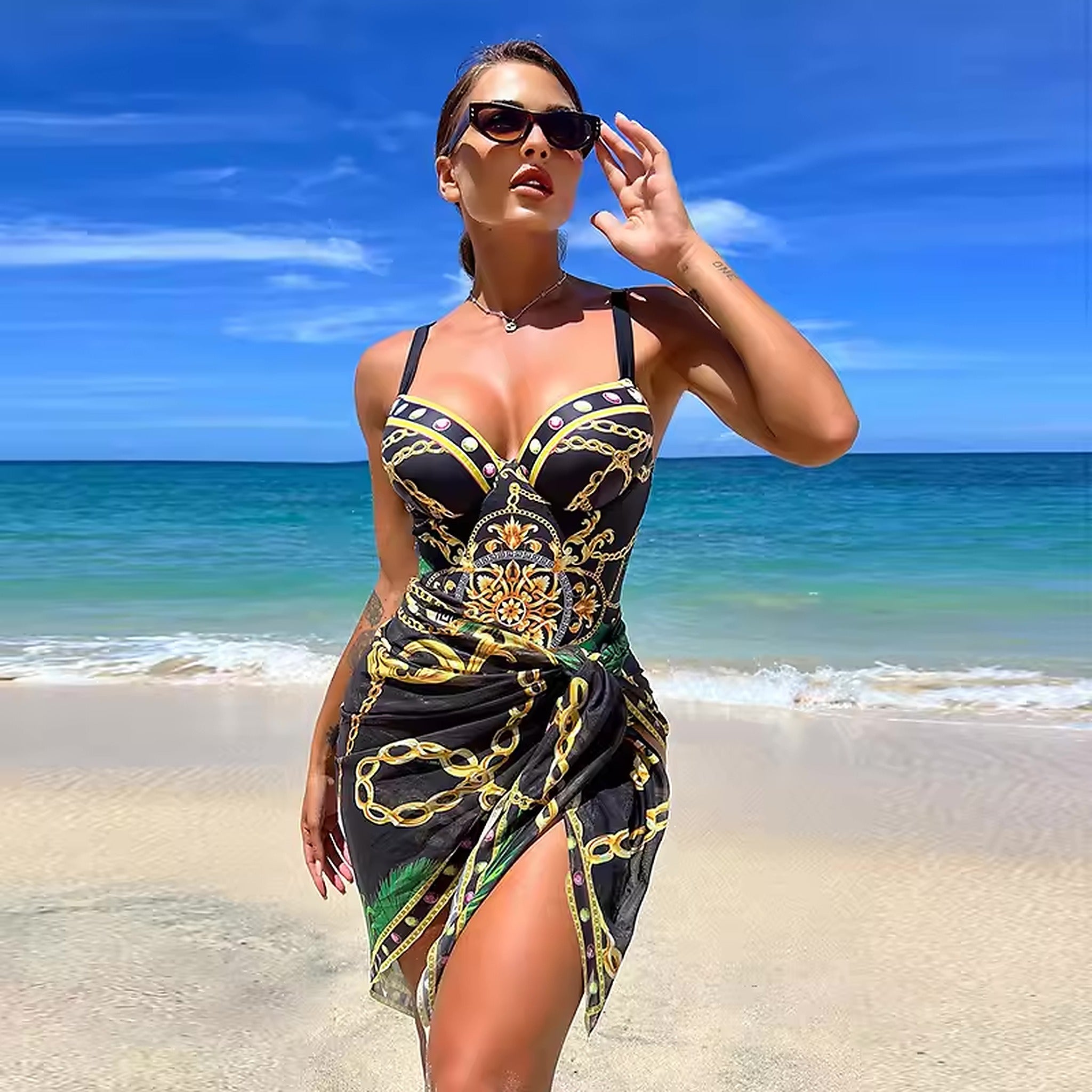 Woman wearing black and gold chain-print bikini with matching sarong on beach with turquoise ocean and blue sky.